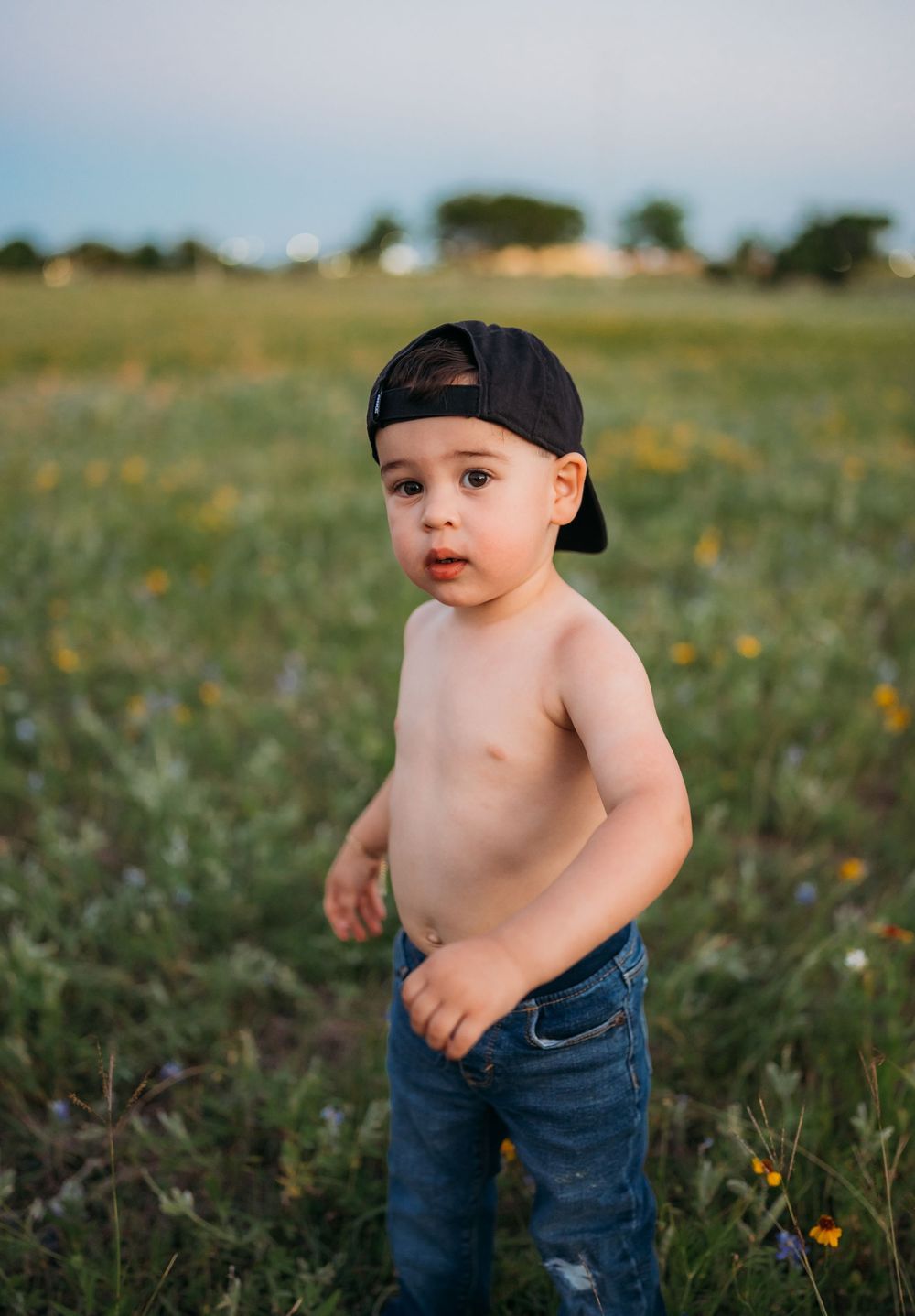 Toddler wearing backwards baseball cap and jeans stands shirtless in meadow with wildflowers at dusk.