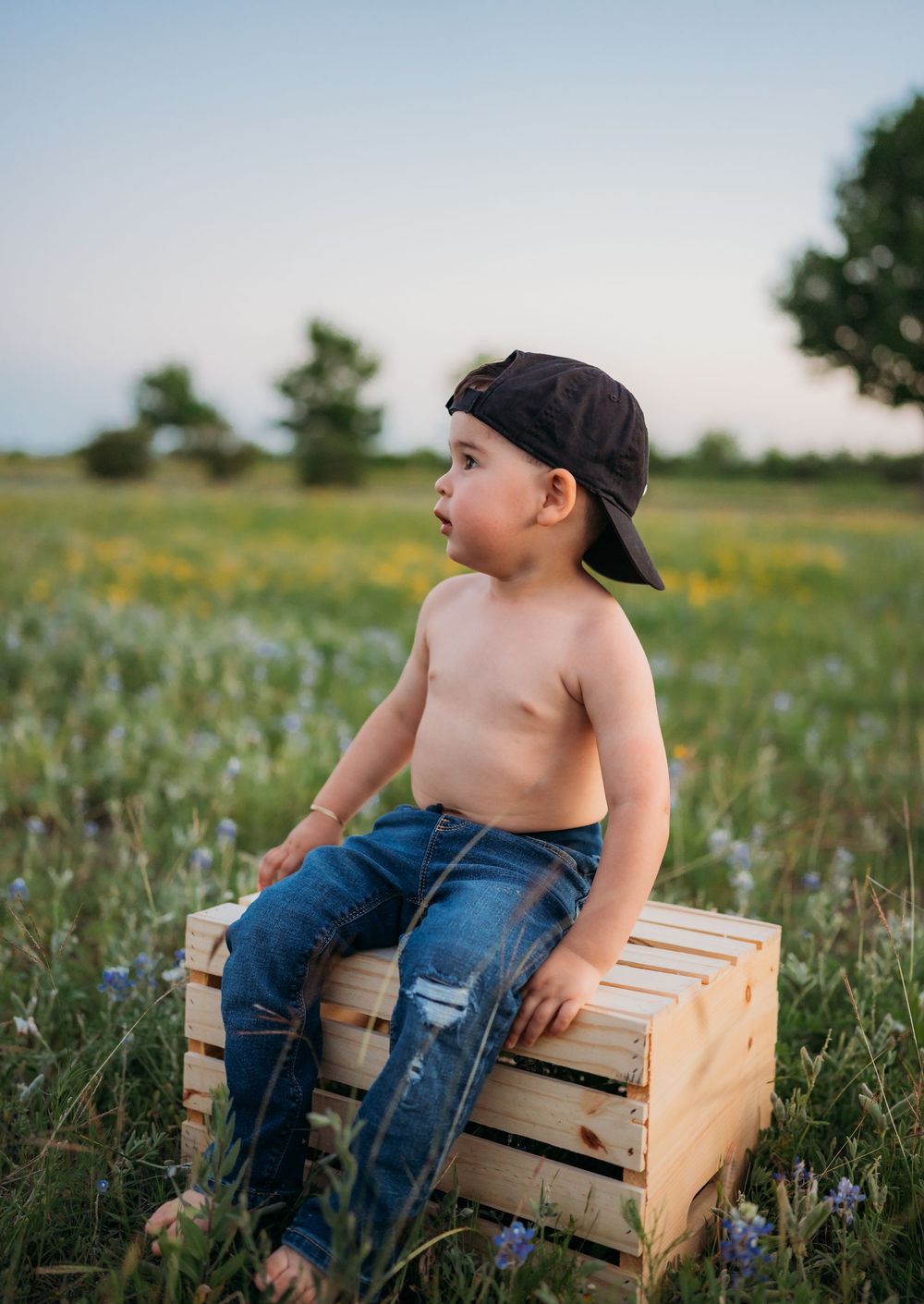 A shirtless child in ripped jeans and backwards cap sits on a wooden crate in a wildflower meadow at sunset.