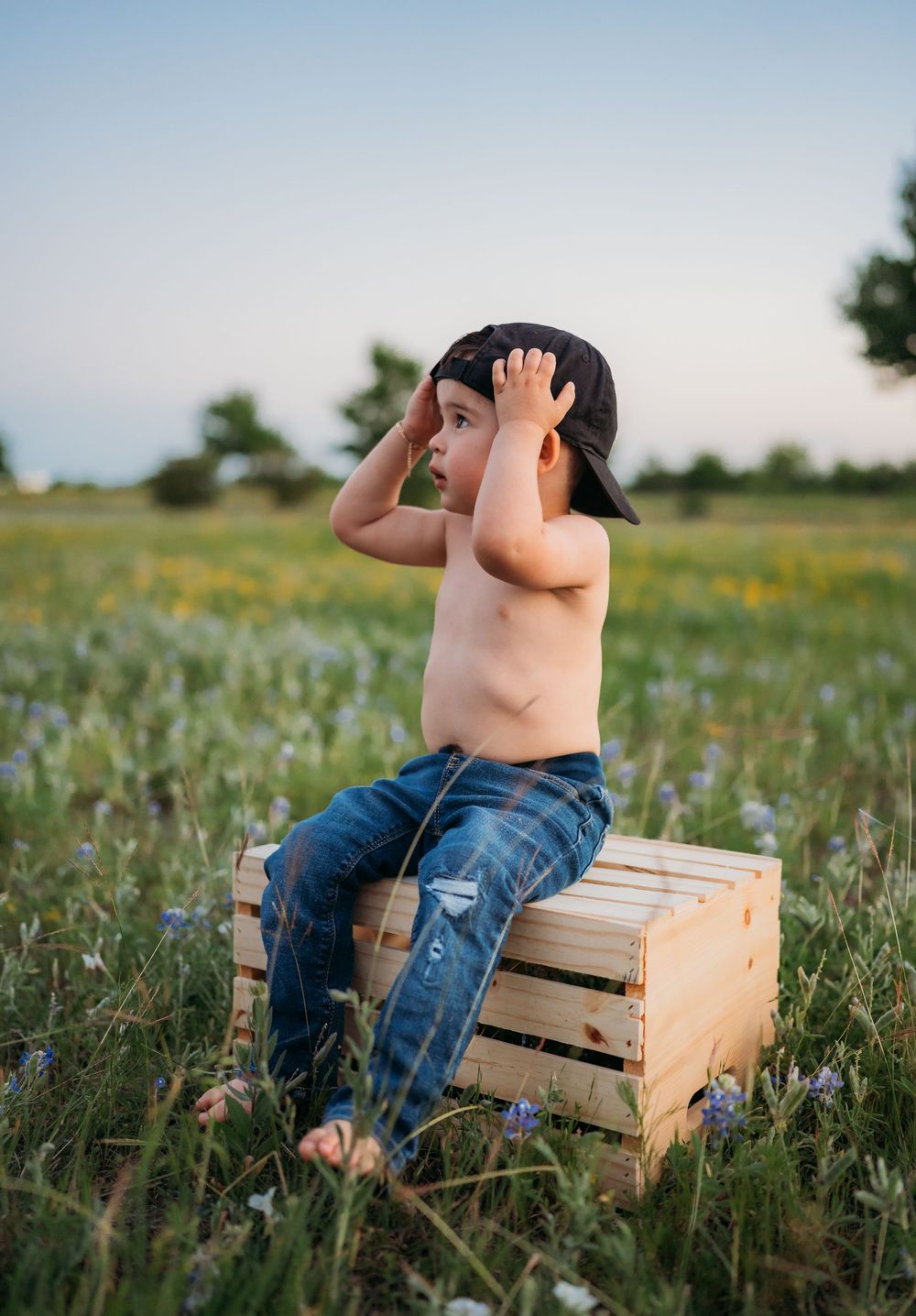 Small child in jeans sits shirtless on wooden crate in wildflower meadow at sunset.