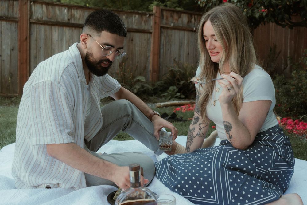 A couple sits on a white blanket having an intimate conversation in a backyard setting.