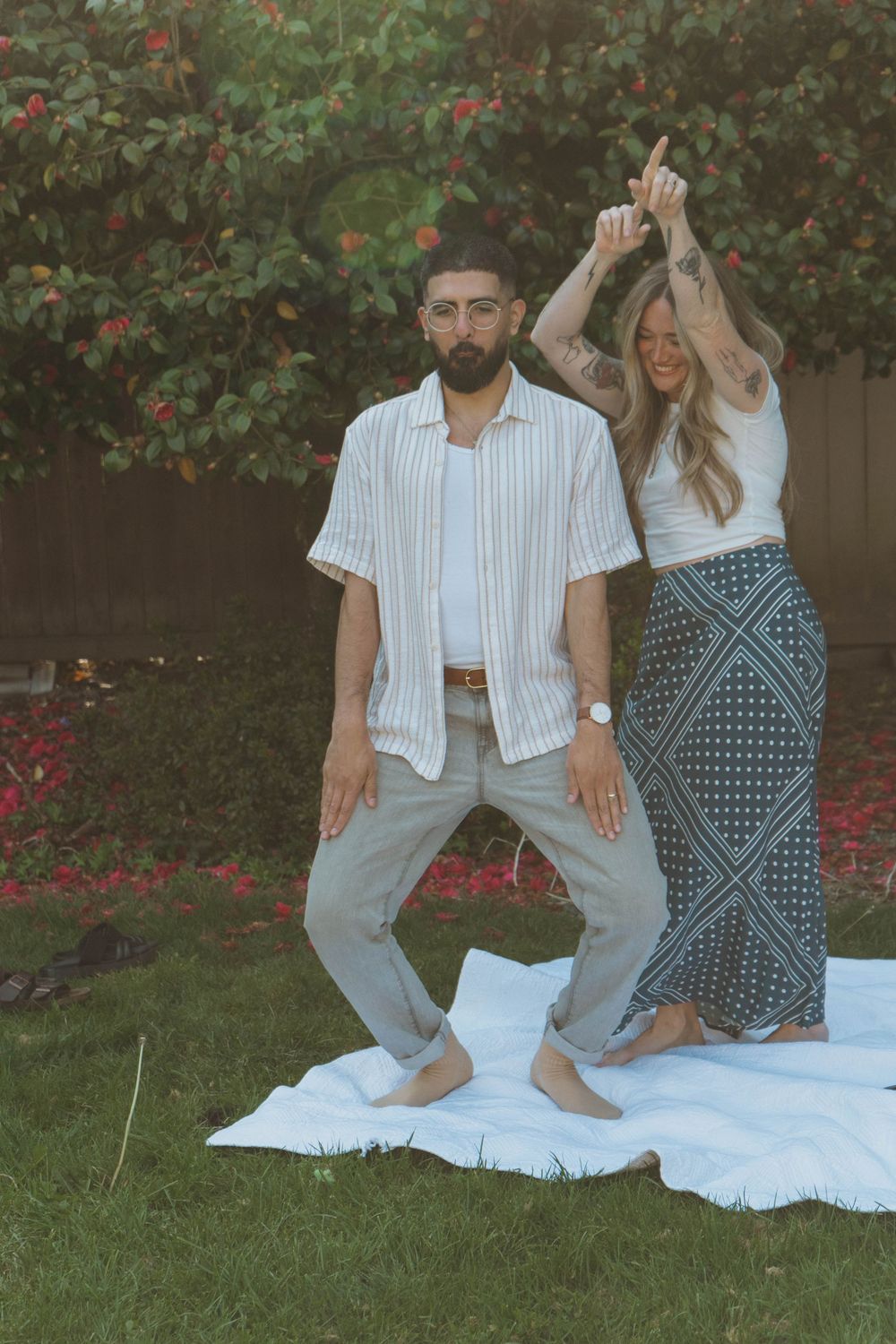A couple enjoys a casual photoshoot in a garden with red flowers on a white blanket.