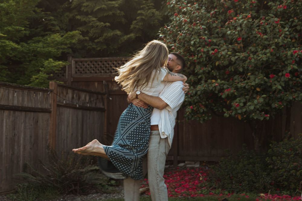 A couple shares a romantic moment in a garden while she lifts her leg during their embrace.