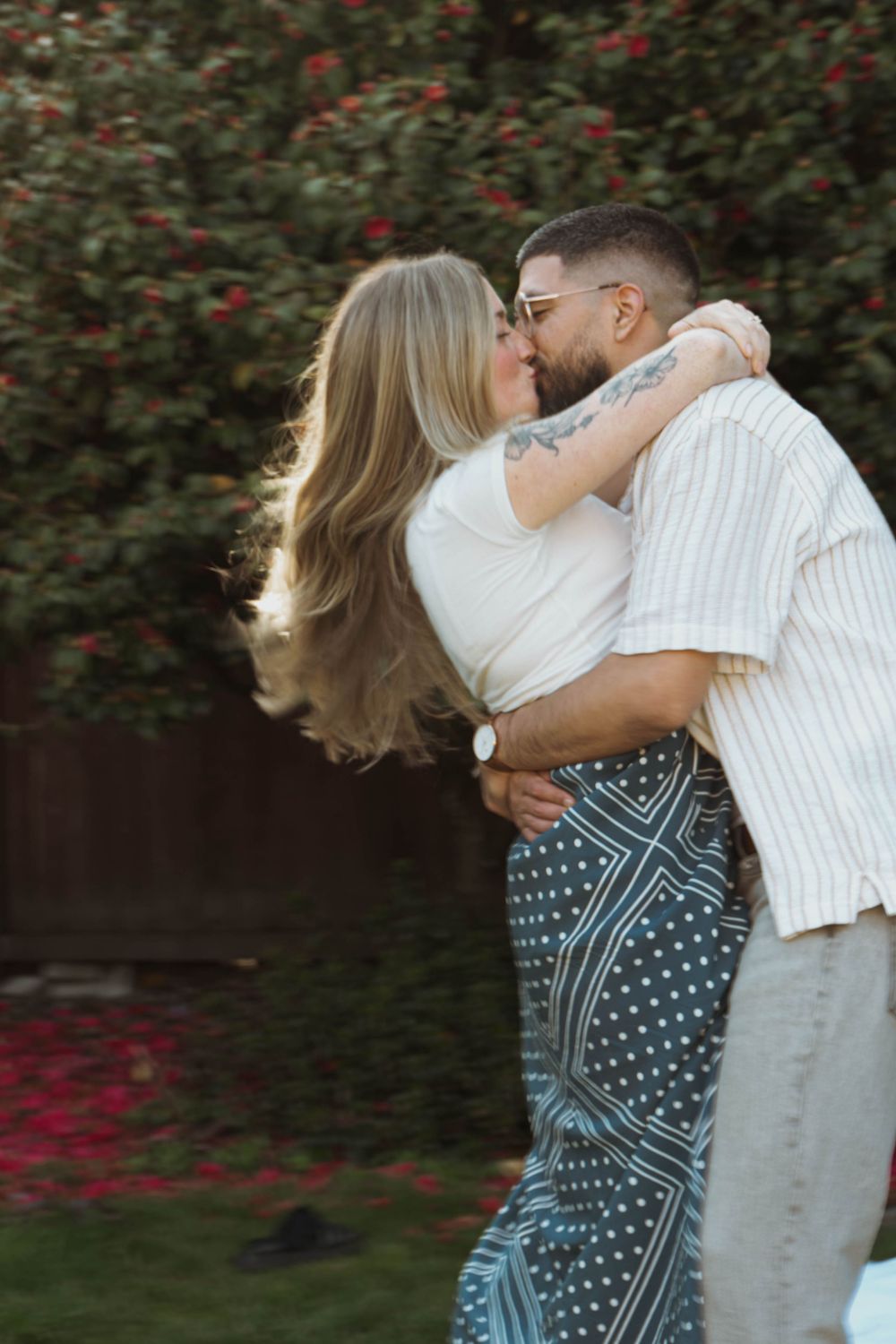 A romantic couple embraces and kisses in front of blooming red flowers and greenery.