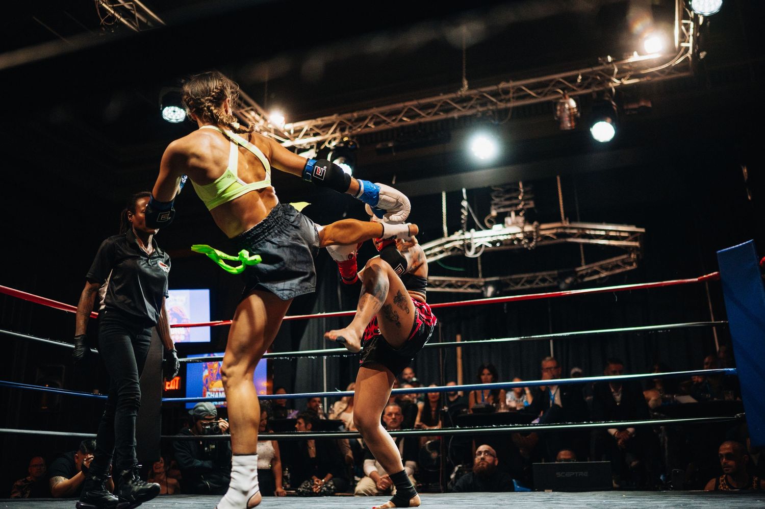 Two Muay Thai fighters exchange powerful kicks during an intense match in a boxing ring under bright spotlights.