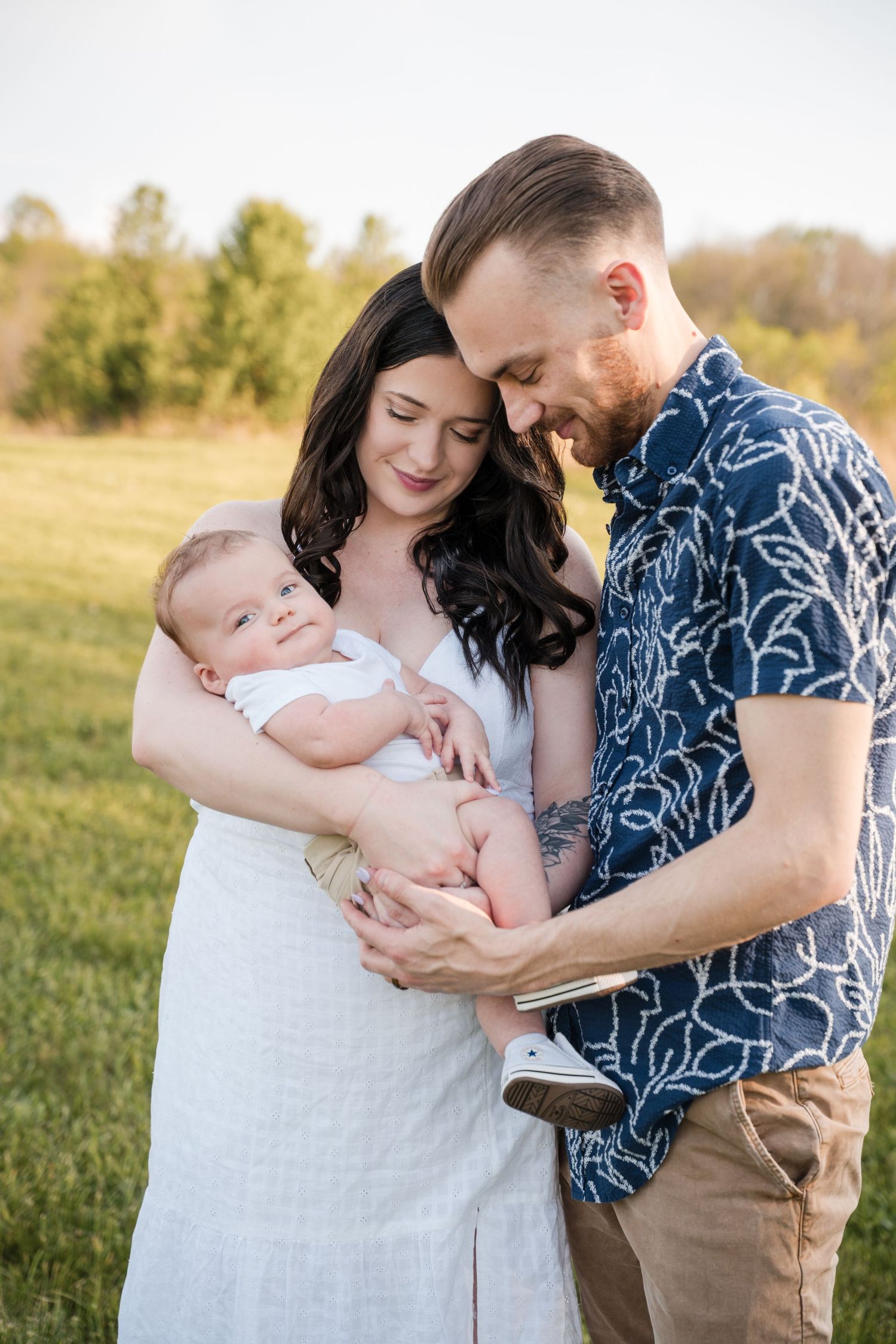 A tender family moment outdoors as new parents hold their baby in a golden grassy field during sunset.