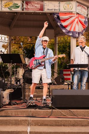Musician in blue shirt and shorts plays guitar on outdoor stage decorated with patriotic bunting.