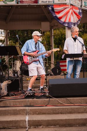 Musicians perform on elevated outdoor stage with patriotic decorations and sound equipment.