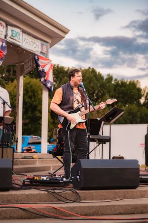 Guitarist in dark vest performs on outdoor stage at sunset during live concert.