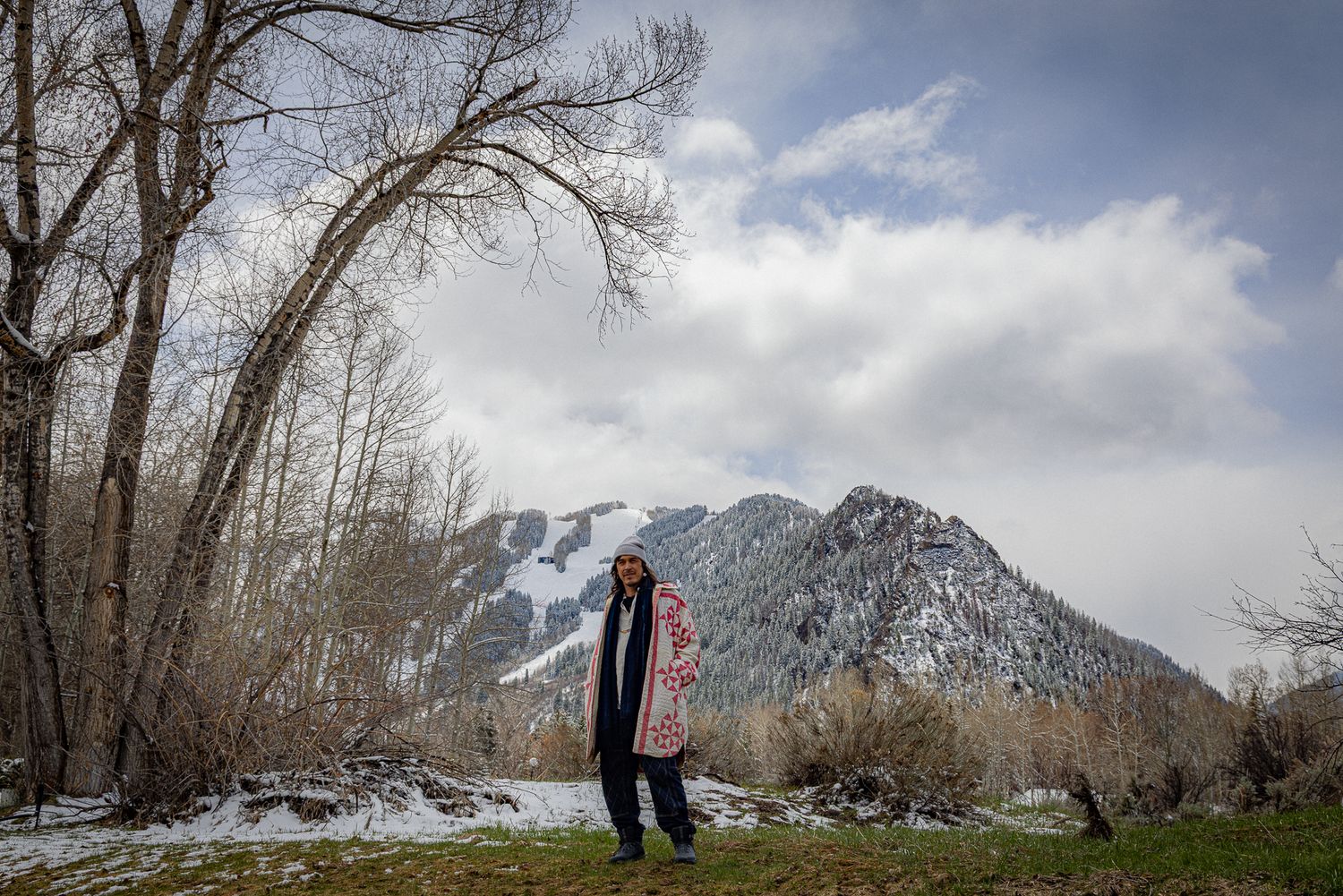 A winter scene of someone standing in snow near bare trees with rugged mountain peaks and cloudy sky in the background.