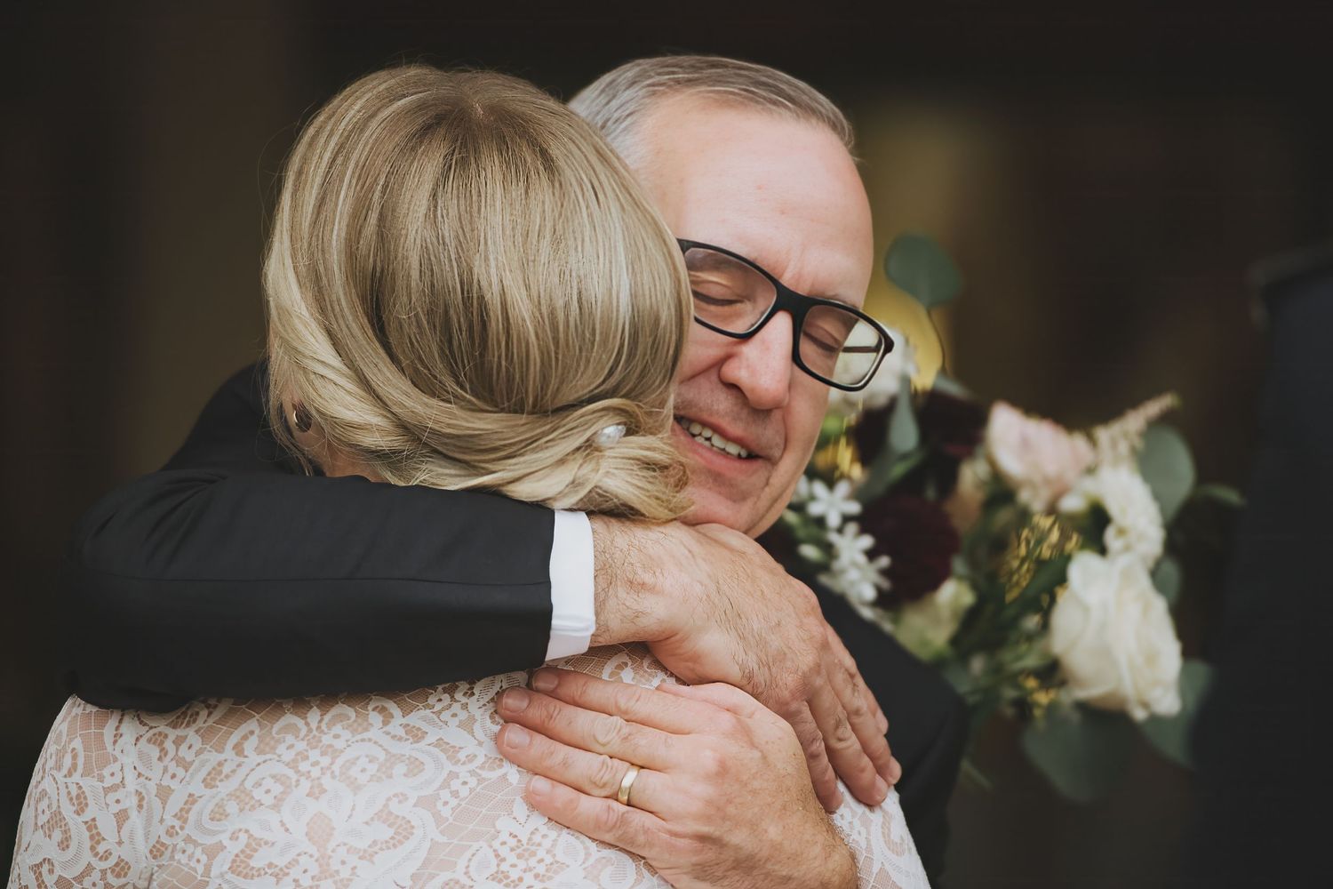 A tender emotional embrace between father and bride at a wedding ceremony with a bouquet of white flowers visible in the background.