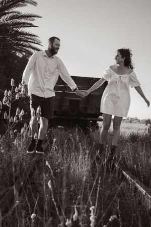Couple in white clothing walks through a field of tall grass at sunset.