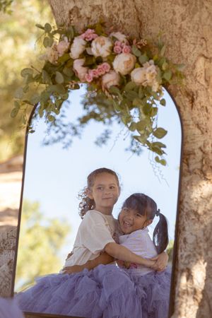 Children in white and purple dresses play under floral archway in sunlit garden.
