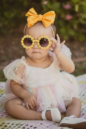 Baby girl in white tulle dress wearing yellow daisy sunglasses and bow headband sitting outdoors.