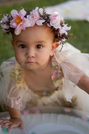 Baby girl in floral crown and tulle dress poses outdoors on grass during first birthday cake smash photo session.