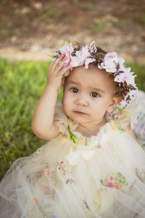 Adorable baby girl in floral tutu dress and pink flower crown sitting outdoors on green grass.