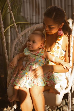 Young mother in floral bikini holds baby in matching dress on macrame swing chair in golden sunlight.