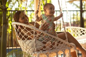 Mother and baby relaxing together in a macrame hanging chair on a tropical balcony surrounded by palm trees.