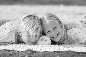Two smiling blonde children lying on a blanket with a small white guinea pig in a black and white photo.