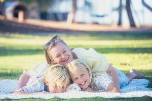 A joyful mother and two blonde children laughing together on a blanket in a sunny green park.