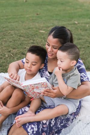 Family members lying on a blanket outdoors while reading a book together.
