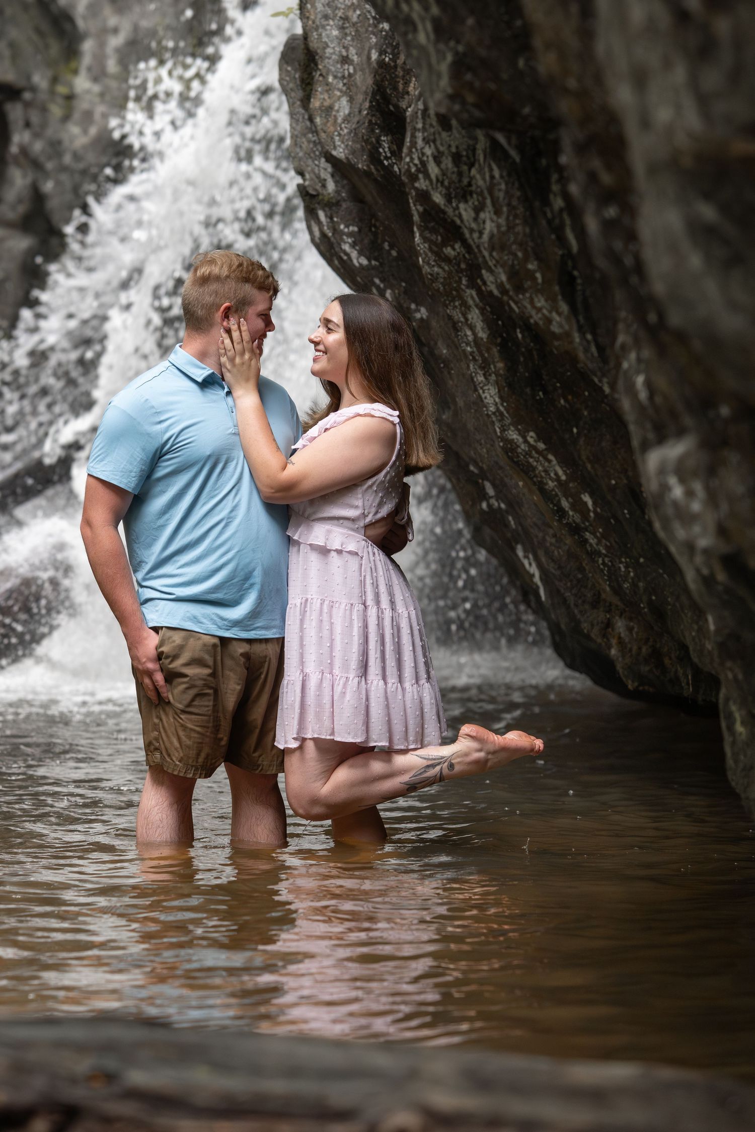 A couple stands in shallow water near a dramatic waterfall during an engagement session.