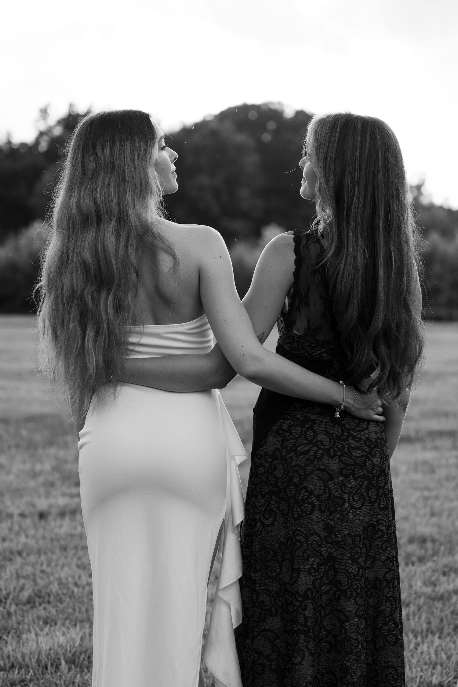 Two sisters in formal dresses embrace while standing in a field at sunset.