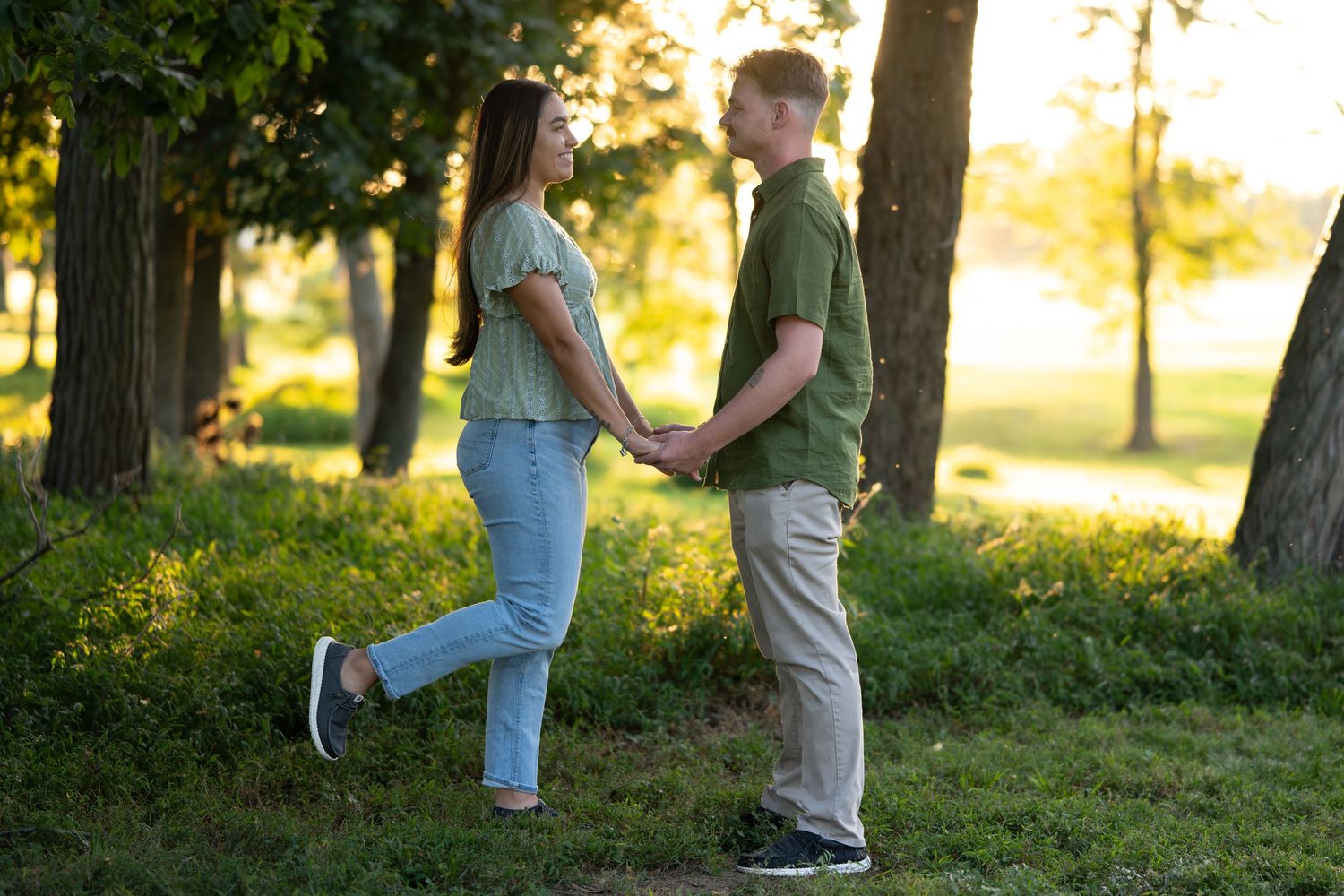 Couple holds hands while standing in a sunlit park with trees in the background.