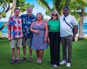 A group of five people pose together on a lush tropical lawn with palm trees and greenery in the background.