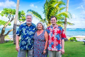 Three people wearing colorful Hawaiian shirts and summer attire pose together on a tropical beach with palm trees and ocean.