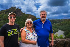 Three people pose together on a hiking trail with dramatic mountain scenery and blue cloudy skies in the background.