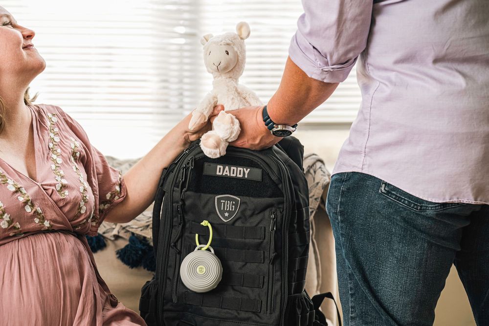 A black tactical diaper bag backpack sits between two people wearing casual clothing in a home setting.