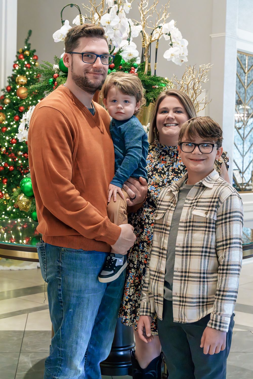 A family of four poses together by a decorated Christmas tree and white orchids in an elegant indoor setting.
