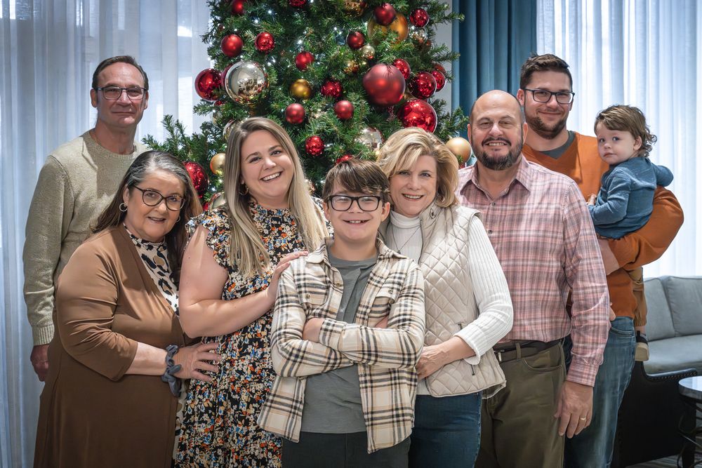 A group of people pose together in front of a decorated Christmas tree with red ornaments in a living room setting.