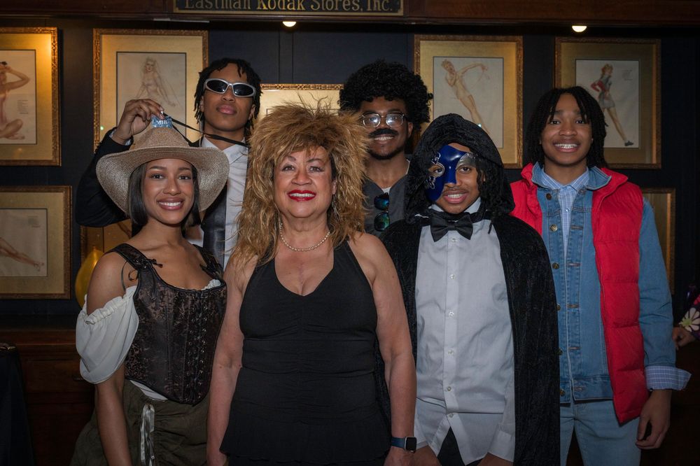 A group of people dressed in various costumes pose together in front of framed artwork on a wall.
