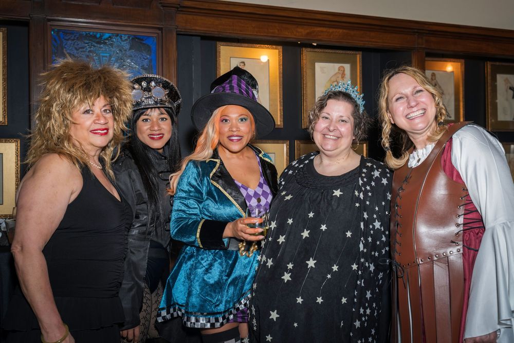 A group of friends in festive party attire gather together at an indoor event for Halloween.