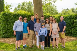 A group of people in coordinating casual attire pose together outdoors in front of a manicured green hedge on a sunny day.