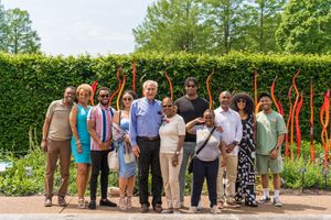 A group of people pose together outdoors in a garden with tall orange sculptures and green hedges in the background.