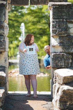 Couple sharing a playful moment at stone archway near water fountain in park on sunny day.