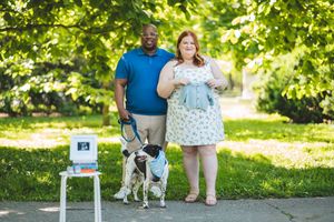 Couple standing with their dog in a sunny outdoor park setting next to a small table with decorative items.