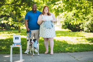 Couple standing outdoors with their dalmatian dog and ultrasound photo displayed on small decorative table in a sunny park.