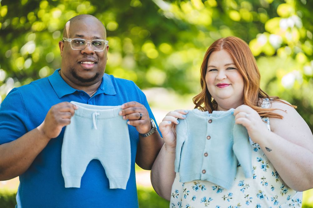 Smiling couple outdoors holds small blue baby clothes while standing together in a garden setting.