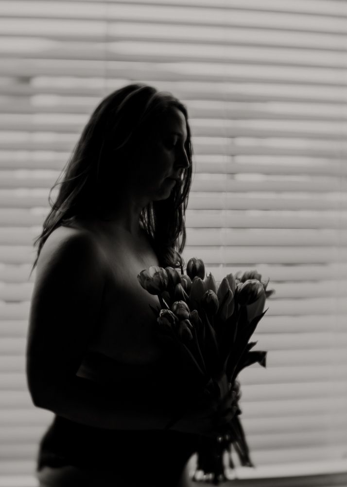 Silhouette holding bouquet of flowers against venetian blinds in monochrome photography.