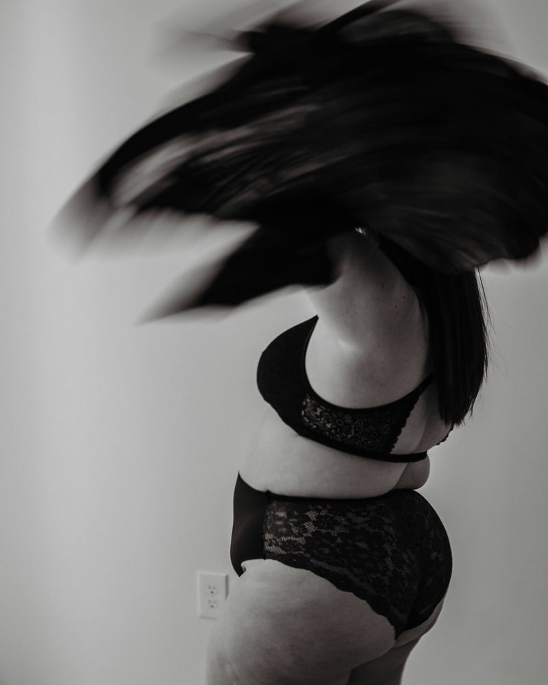 Black and white artistic boudoir photo showing silhouette in dark lingerie with dynamic hair movement.