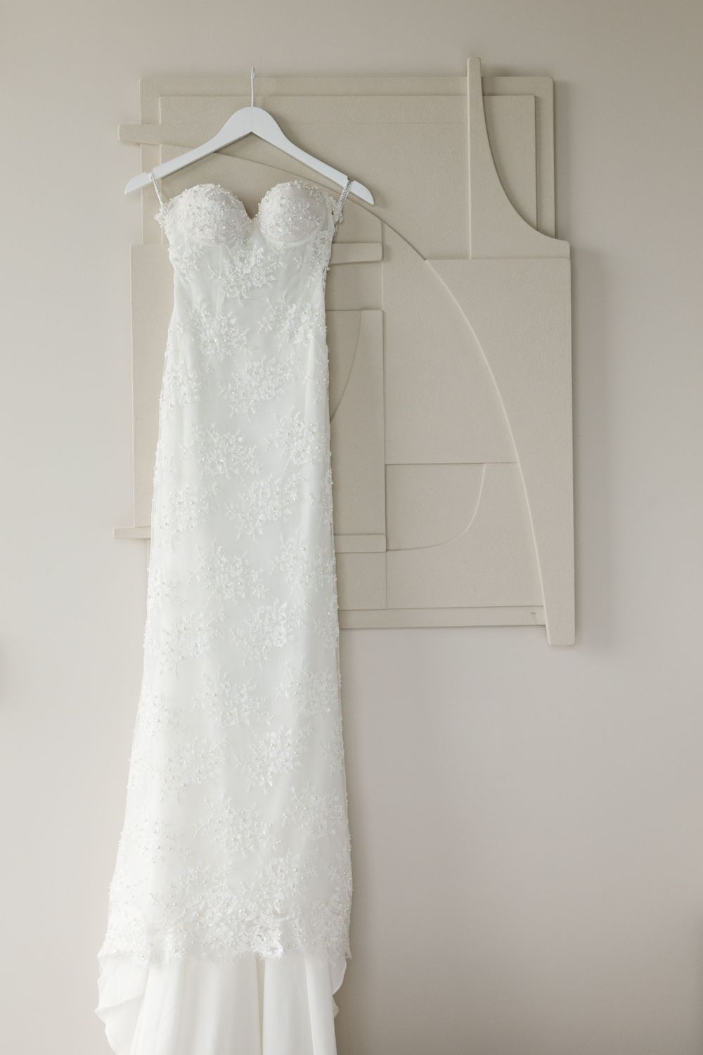 A white lace strapless wedding dress hangs elegantly on a hanger against a neutral wall with modern artwork.