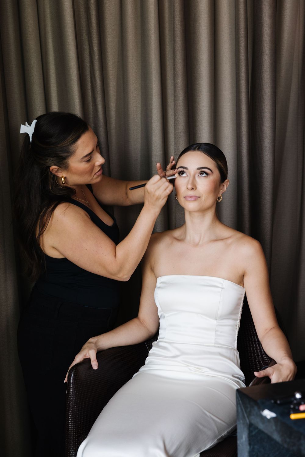 Makeup artist applying eye makeup to bride wearing strapless white dress during wedding preparation.