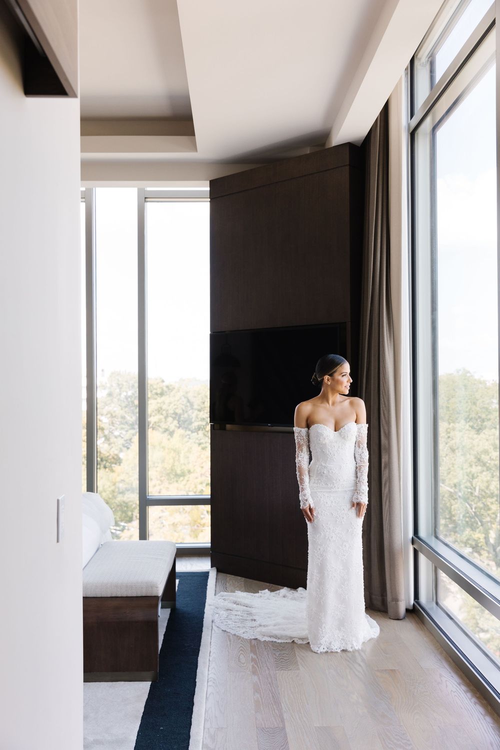 Elegant person in a white lace off-shoulder wedding gown stands by floor-to-ceiling windows in a modern light-filled room.