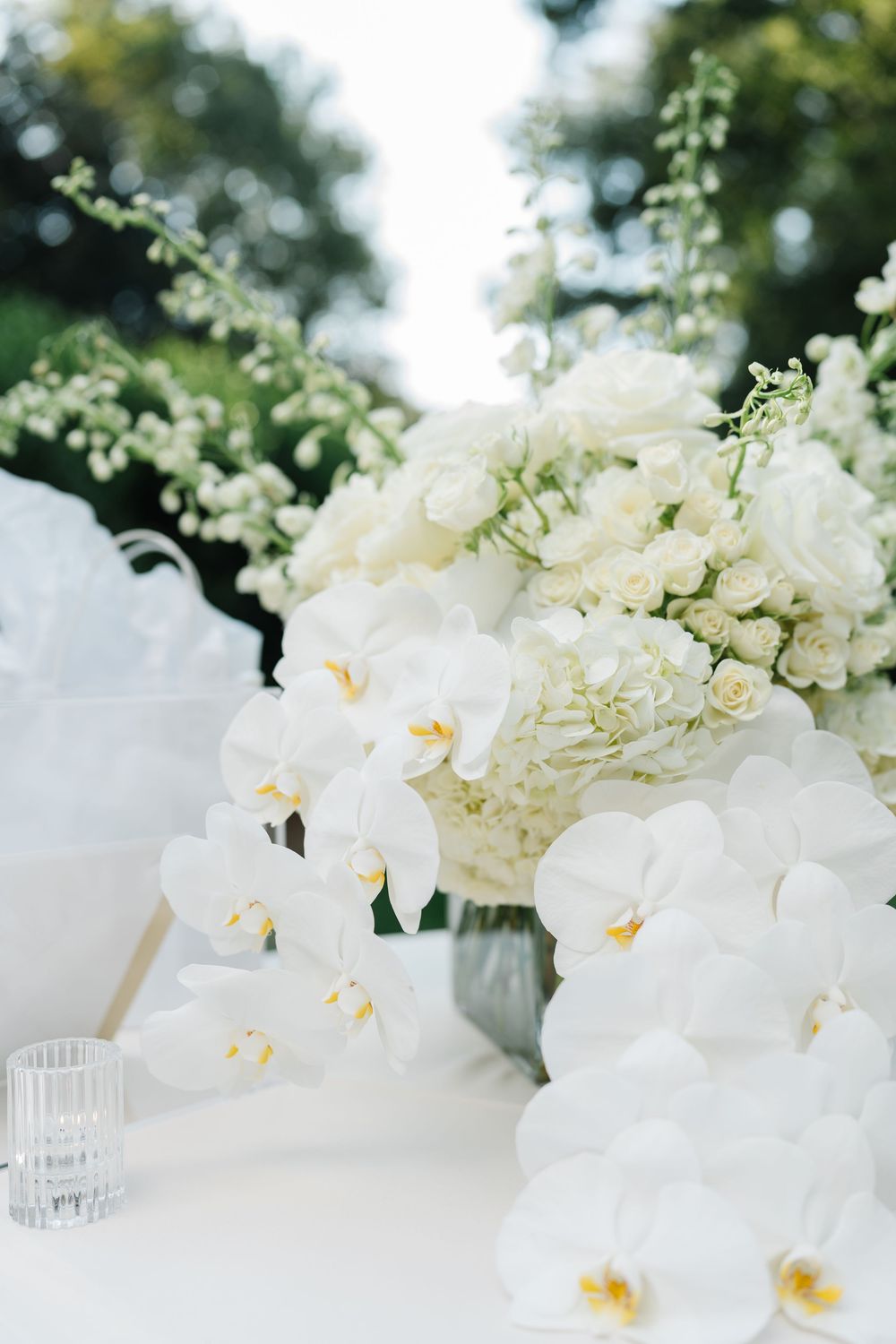 An elegant white floral arrangement with orchids and hydrangeas in a crystal vase on a white tablecloth.