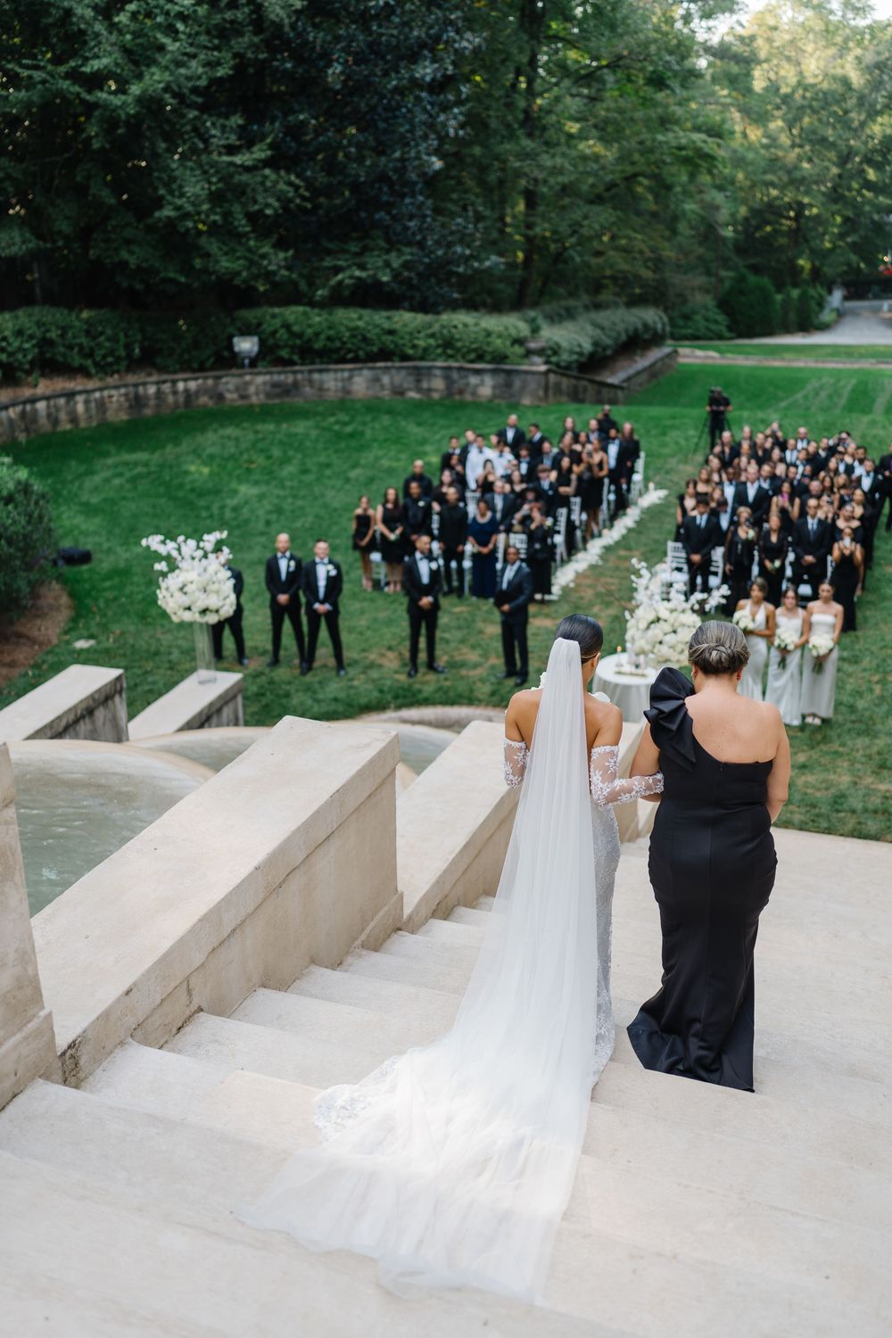 A wedding ceremony with guests gathered in a luxury garden courtyard viewed from behind a bride in white dress and veil on stone steps.