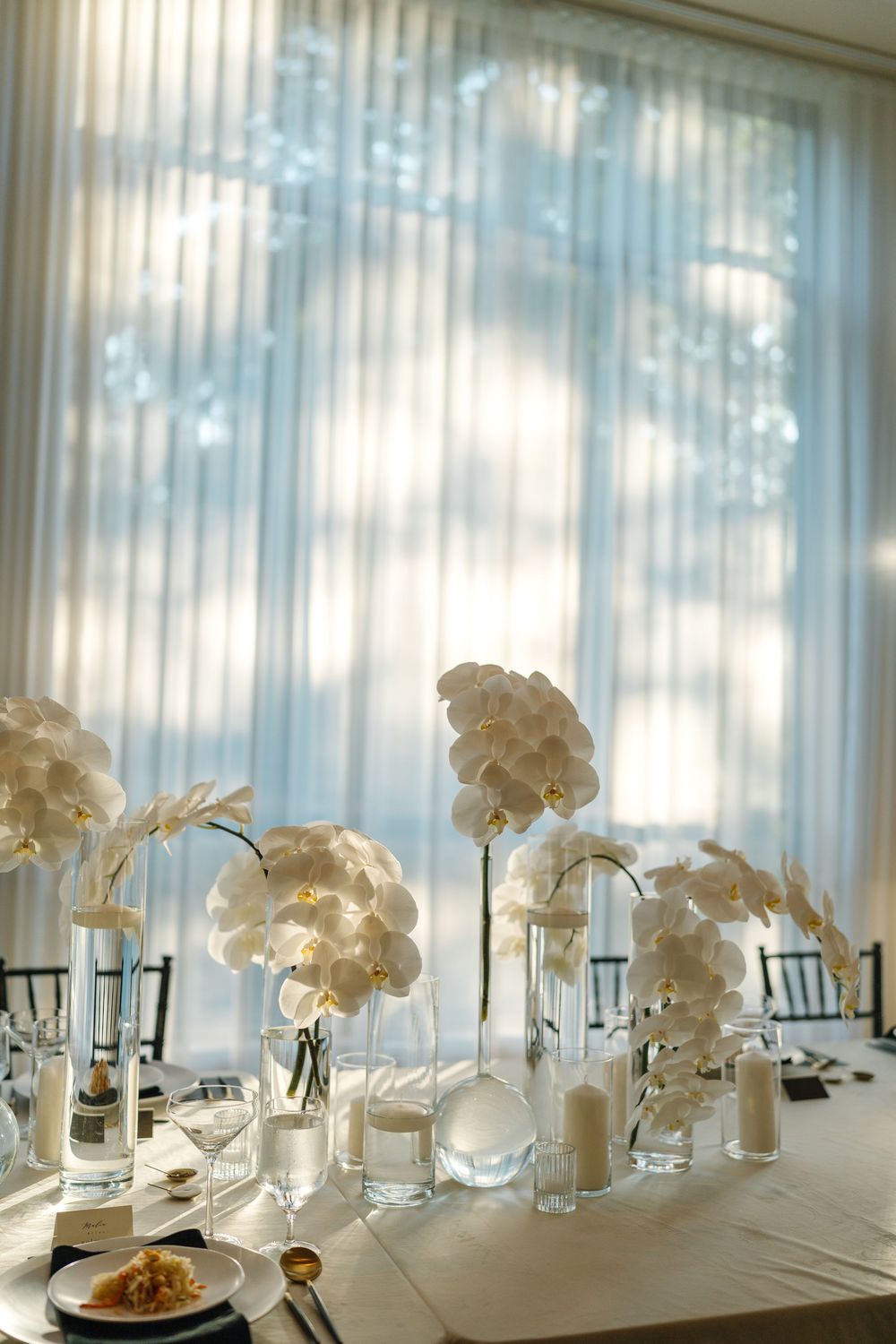 Elegant white orchid centerpieces in glass vases line a formal dining table with soft natural light filtering through sheer curtains.