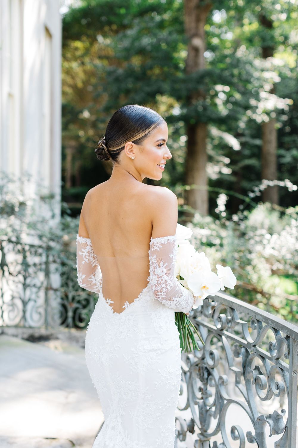 Beautiful backless white lace wedding gown with off-shoulder sleeves shown on outdoor balcony with ornate iron railings.